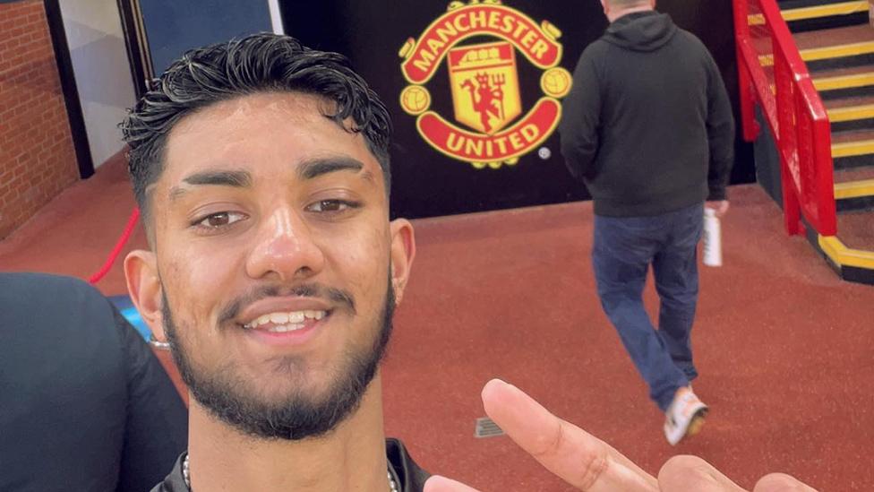 A young man stands in front of a black board with the Manchester United club crest on it. He's taking a selfie, holding the camera slightly above head height and pointed down towards him. His hair is short and styled in neat curls. The club crest is a yellow shield with a red devil on it, surrounded by banners with the team's name written along the top and bottom.