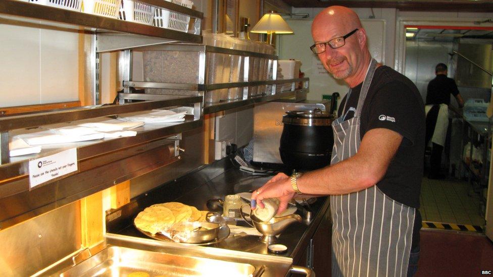 Peter Gibbs preparing food in a silver kitchen. Food lies in large dishes in the foreground. There is a man in the background washing dishes.