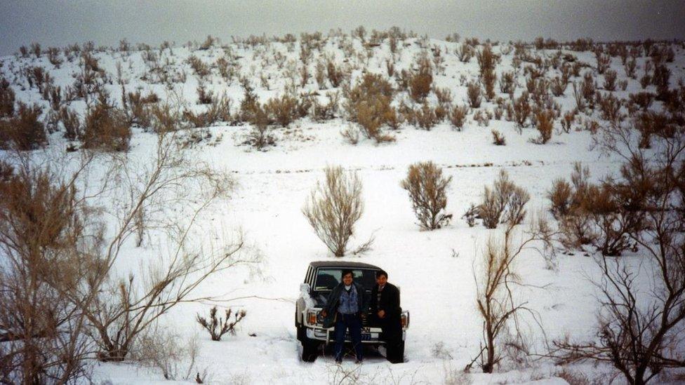 Two people stand in front of a parked car in what appears to be a snow-covered desert.