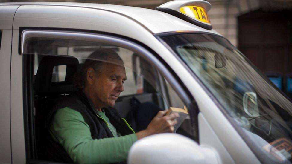 Cab driver Andy Biggs reads a book in a black cab as he queues for a fare outside Victoria Station, London