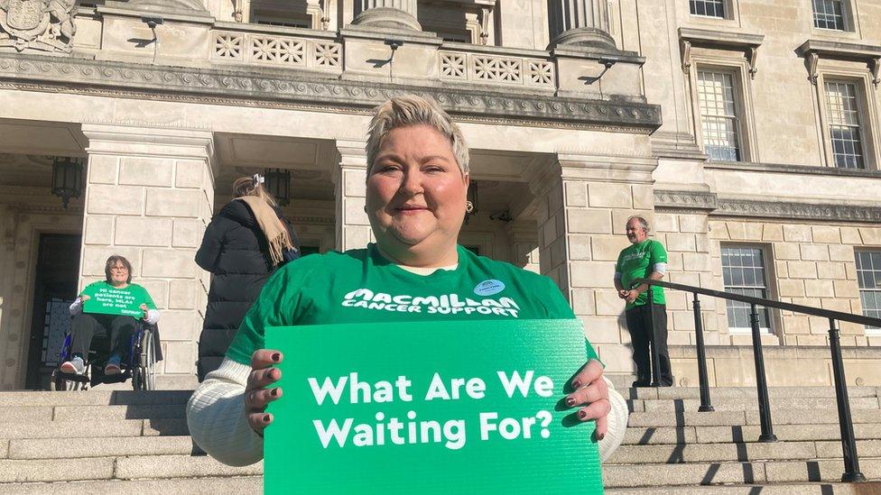 Colleen McCallion standing in front of Stormont holding a sign reading "What are we waiting for?"