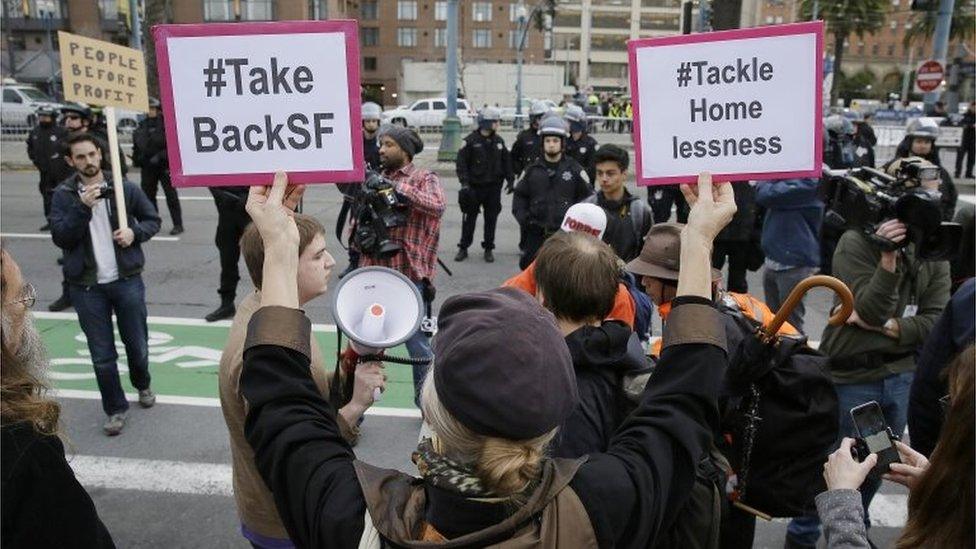 A woman holds up a pair of signs as police look on during a protest to demand city officials do more to help homeless people outside Super Bowl City, a pro-football's weeklong theme park near the famed Ferry Building in San Francisco on 3 February 2016.