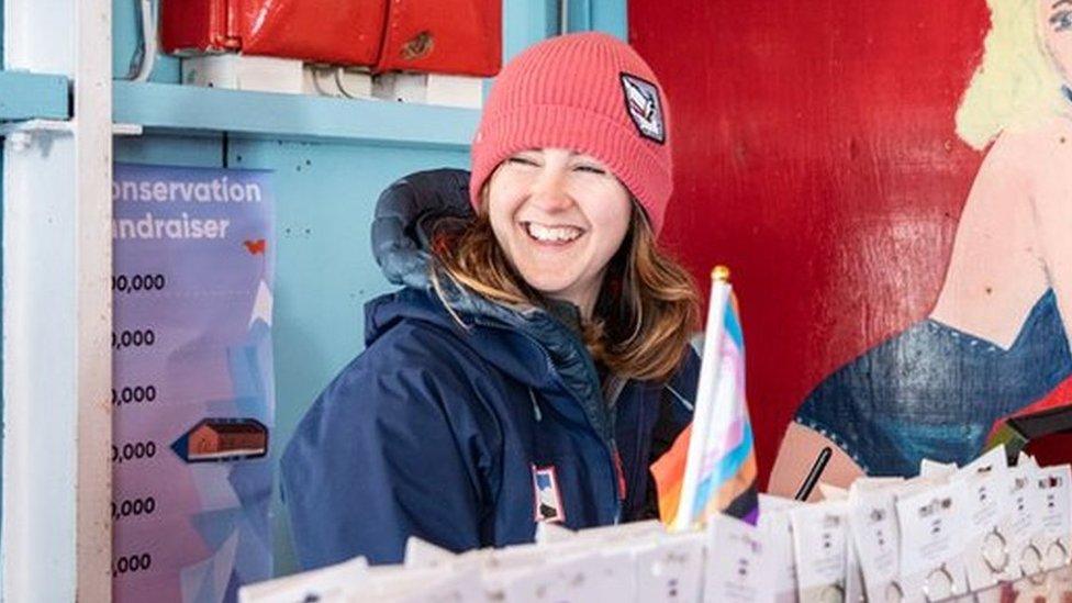 Natalie Corbett smiling inside the post office. She is wearing a navy coat and a red beanie hat. There is a red Post Office sign behind her and key rings on a stand in front of her.