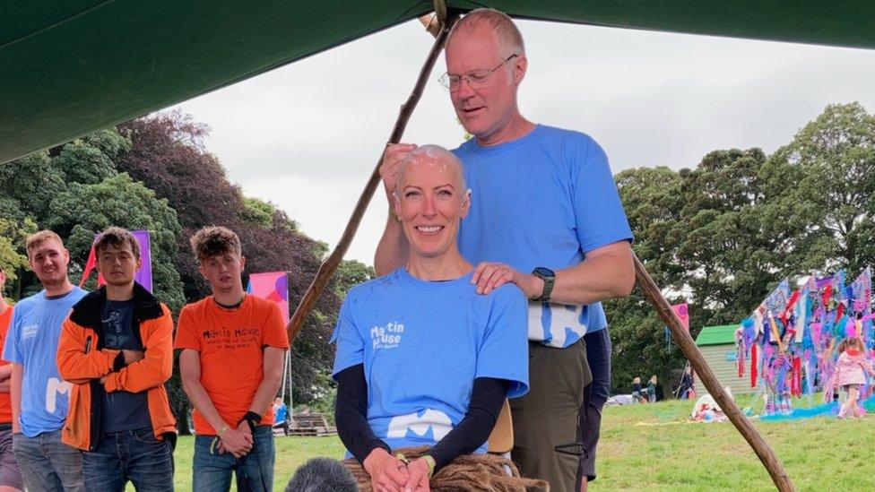 Man shaving woman's head with onlookers behind them