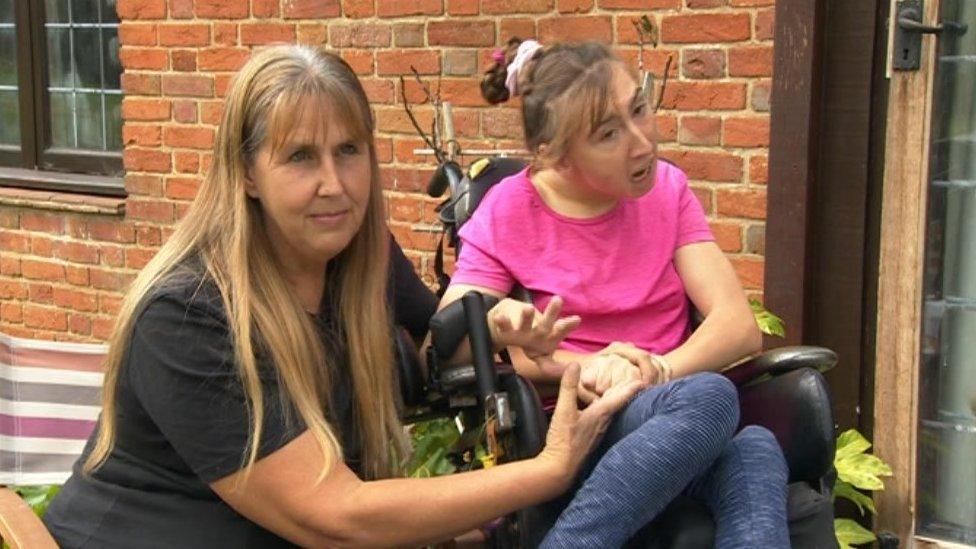 Natasha Lambert (right) sitting in her wheelchair accompanied by her mother Amanda crouching beside her and holding her hand