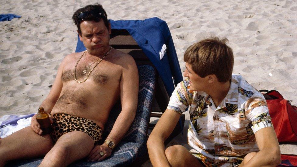 David Jason wearing leopard print swimming trunks and Nicholas Lyndhurst wearing a Hawaiian shirt sitting on sun loungers on a sandy beach