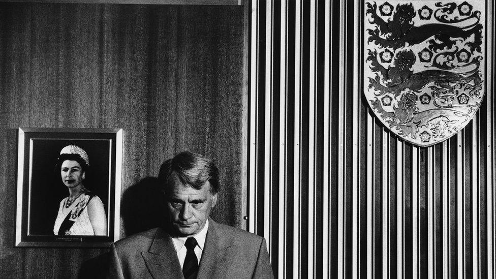 Bobby Robson at a press conference on 24 May 1990 announcing he would leave the England manager's job after the World Cup. Behind him are a photo of the Queen and an England Three Lions crest.