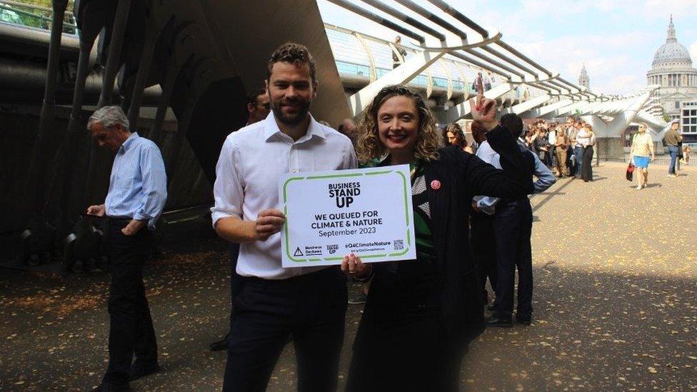 A woman and man hold a sign in front of millennium bridge, saying 'businesses stand up'. She is pointing up. A queue of demonstrators is seen behind them.