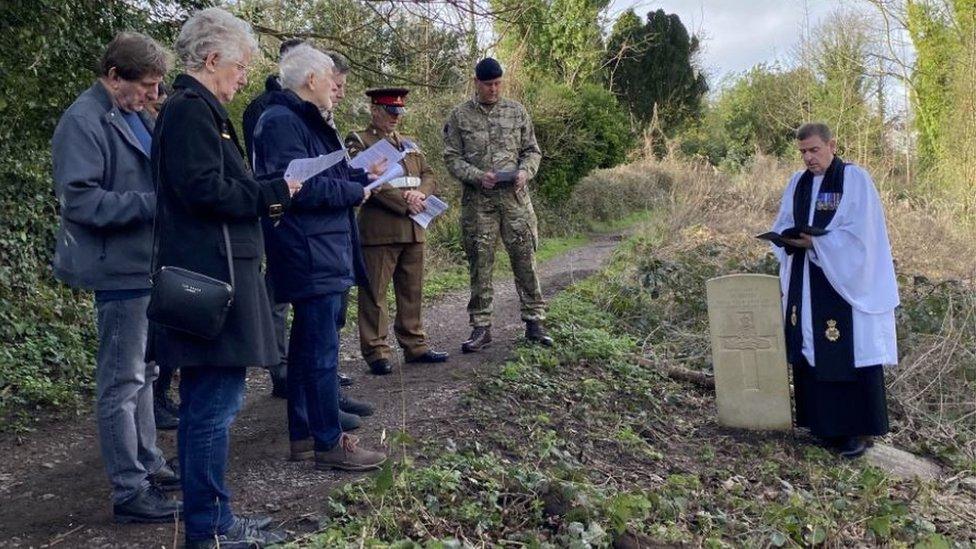 A clergyman reads from a Bible over a grave as uniformed soldiers and a group of people look on