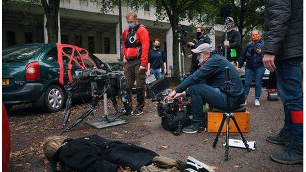 An actor lies on the floor in a chaotic street scene, with the camera operator and crew wearing masks