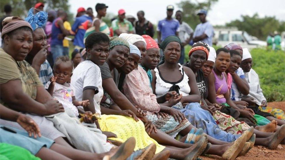 14 women and one baby sitting in a row on the dusty ground