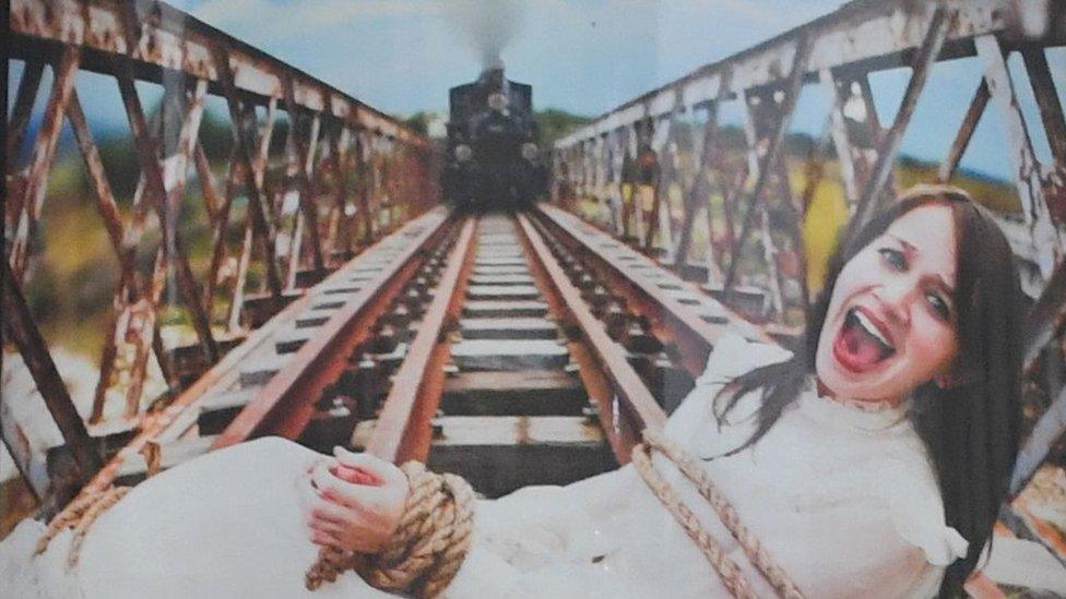 An poster shows a posed shot of a woman screaming at the camera while lying on a railway bridge, bound with rope, while a train approaches