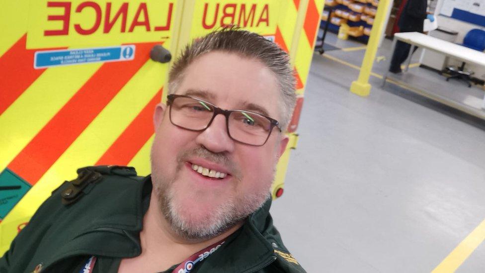 Matt Webb in his East of England Ambulance Service uniform, taking a selfie beside an ambulance at the depot