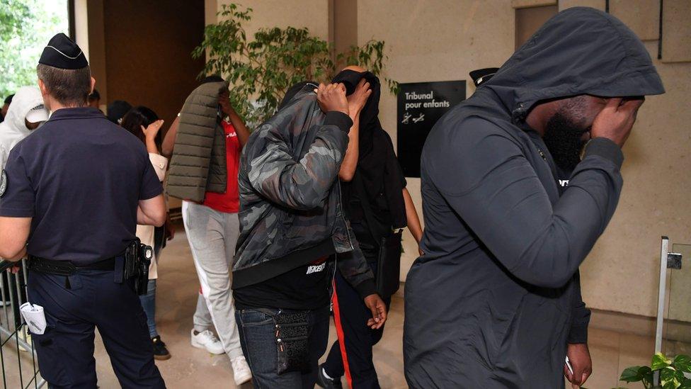 Four man in single file walk past the cameras at a courthouse, obscuring their faces with hoods, jackets and hands