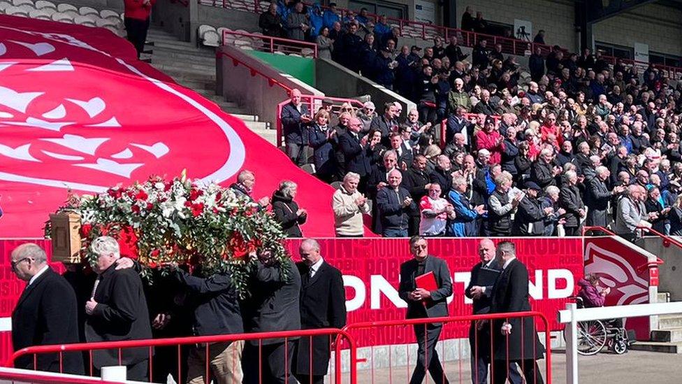 Fans at Craven Park stadium standing up in front of Phil Lowe's coffin