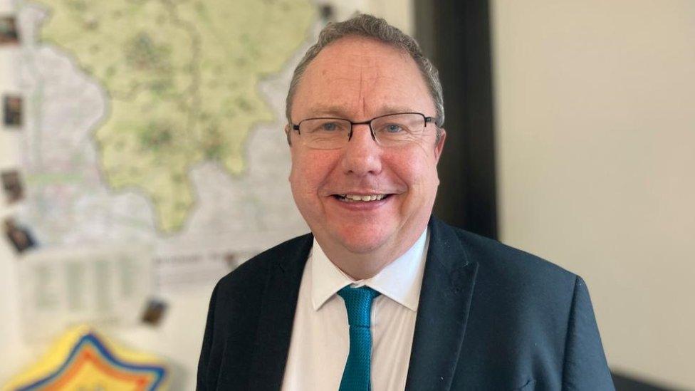 Man with short dark hair and glasses wearing a suit and tie in front of a map