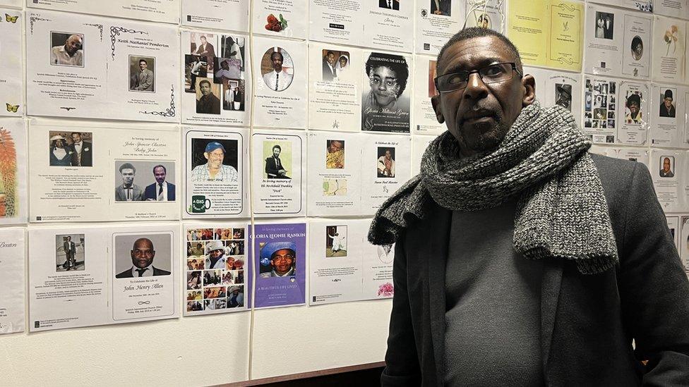 A man stands in from of a wall of pictures of people who have passed away.