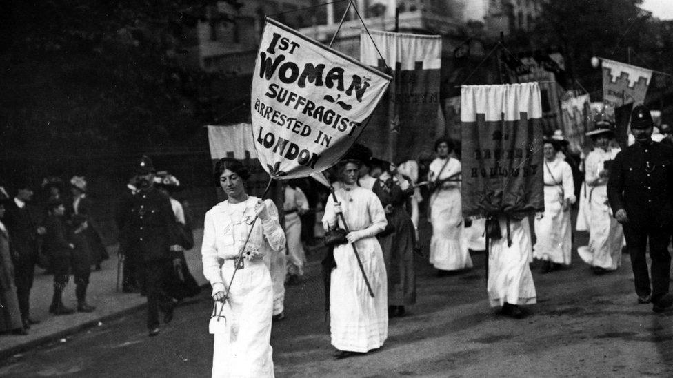 Women wearing long white gowns walk with with placards during a suffragette protest