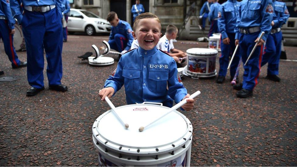 A young boy in a band uniform smiles as he plays a drum