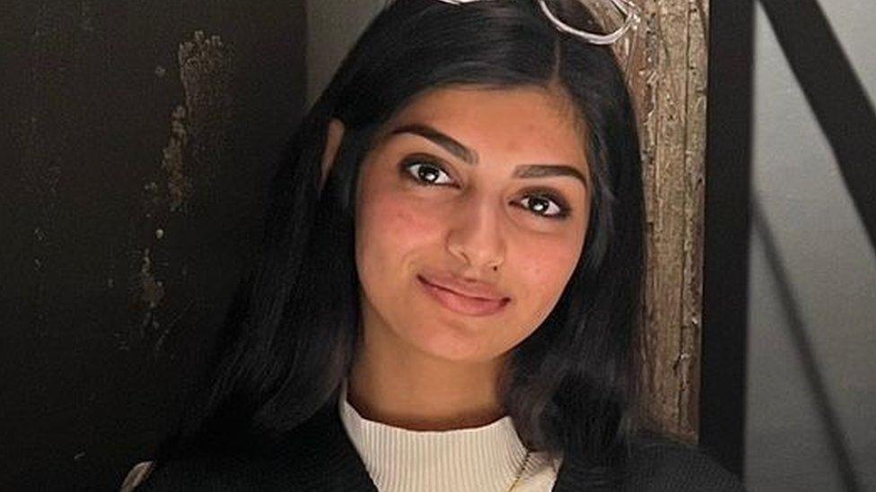 Pria, a young south Asian woman with long dark hair worn loose smiles at the camera. Pria has dark eyes and sculpted eyebrows and wears clear-framed glasses on top of her head. She wears a high-necked top beneath a black woollen cardigan. Pria is photographed inside against a black wall with an exposed wooden feature.