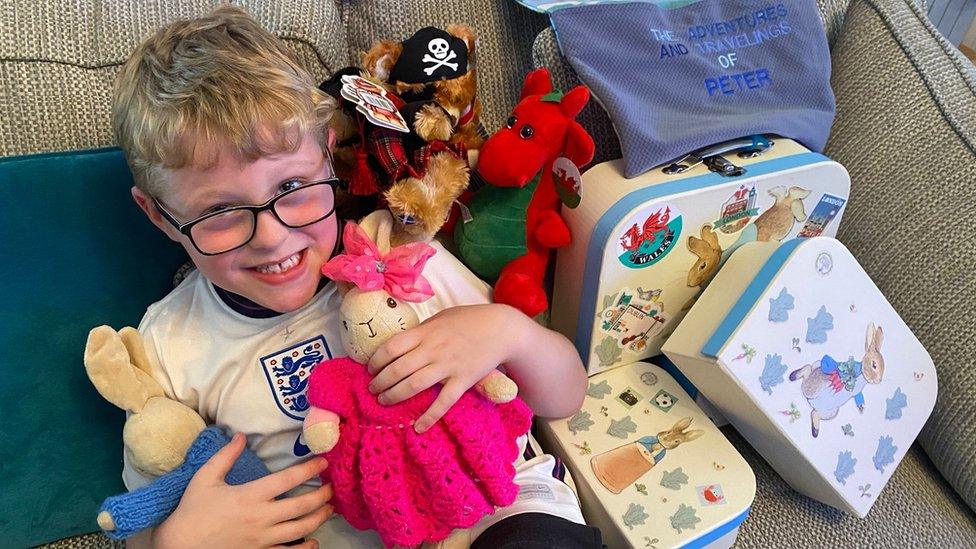 Ethan cuddles Peter Rabbit on sofa surrounded by luggage and postcards