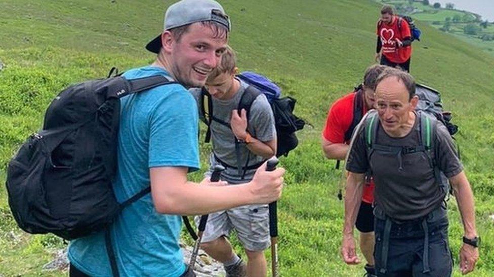 Ben Stewart walking up a hill backwards, looking over his shoulder and using a walking stick