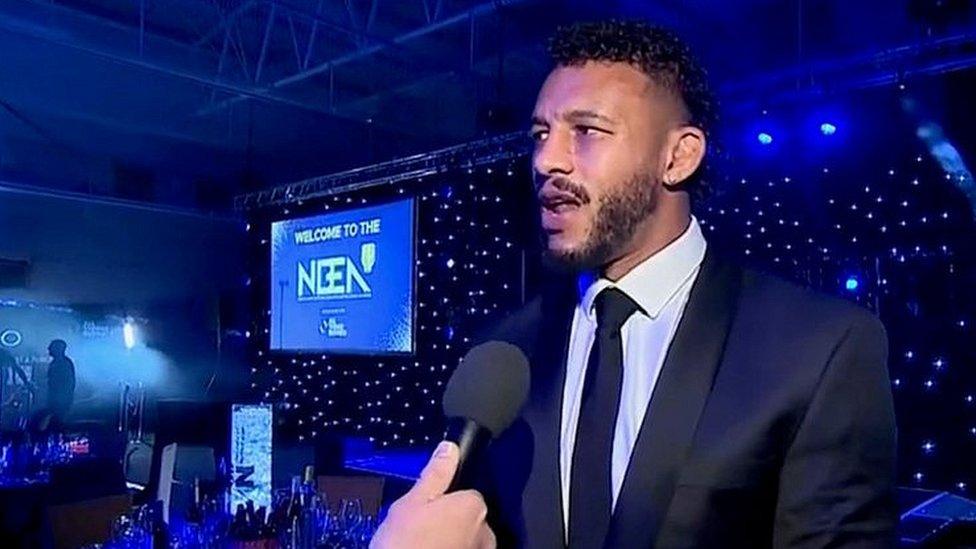 Man with black hair and beard wearing a dark jacket and tie at awards ceremony. He is talking into a microphone.