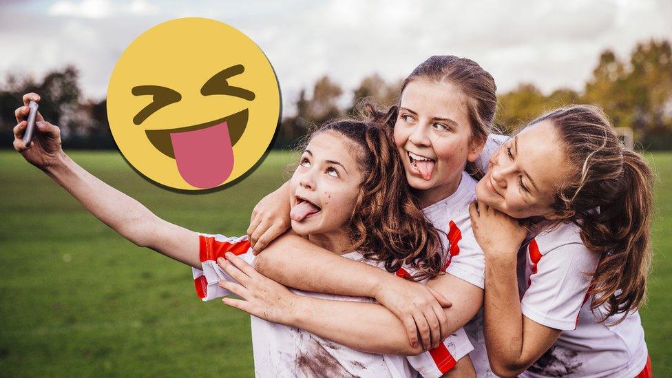 Three girls pull funny faces as they hold up a camera to take a picture and an emoji sticks its tongue out