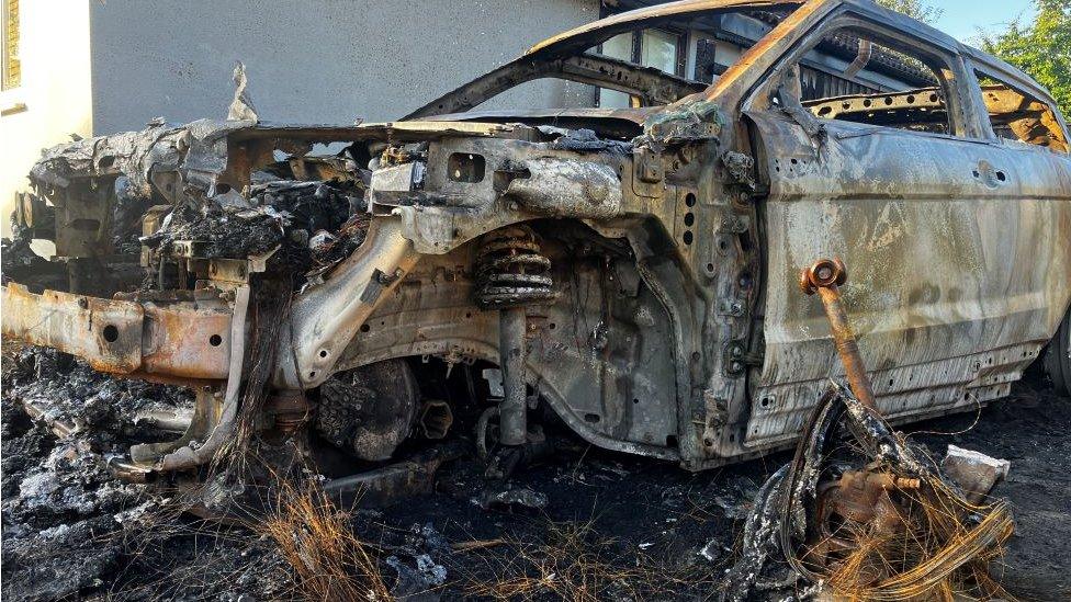 The burnt out remains of a car destroyed in a suspected arson attack. The bonnet is almost entirely gone, parts of the engine are on show an a wheel is missing. A remnant of the wheel is on the ground next to the car shell.