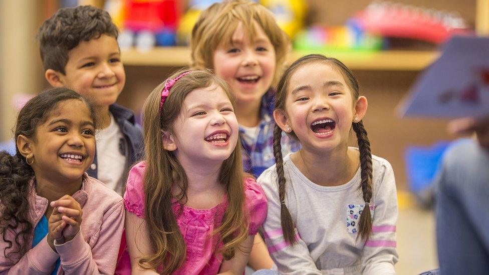 A multi-ethnic group of elementary age children are laughing while their teacher reads them a book during story time