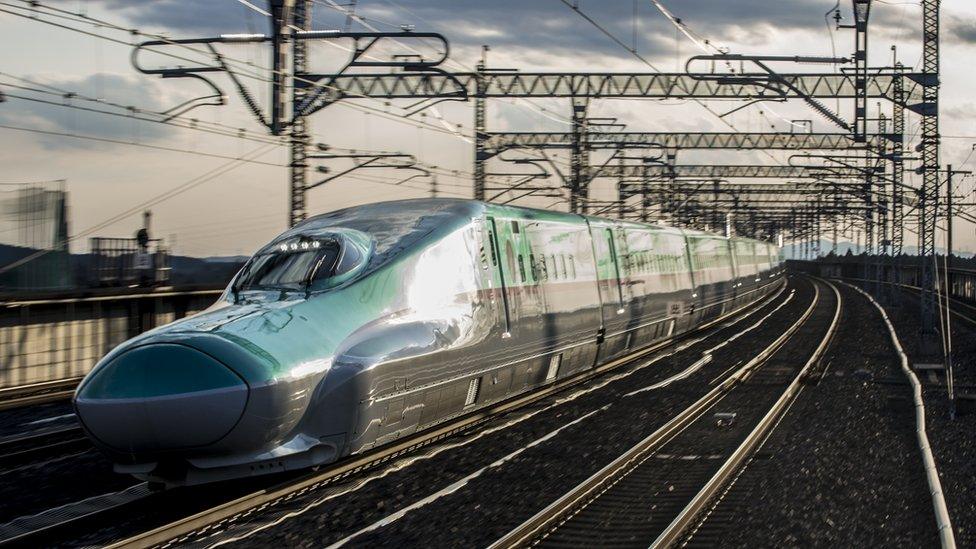 A green bullet train with its long, low nose typical of the type of machine is seen travelling at high speeds against the blurred background of a railway line