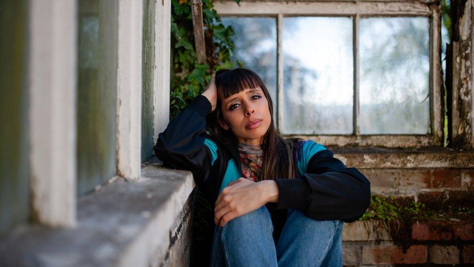 Kayla Painter sitting on the floor with an arm over her knees, leaning her head in her other hand against a wall