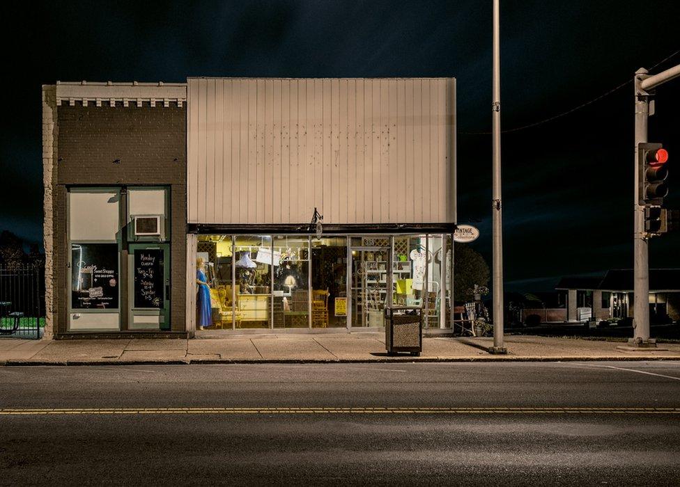 A night view of a closed shop that is illuminated inside, with a mannequin wearing a blue dress