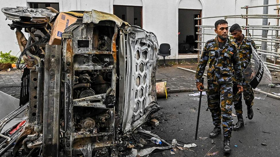 Sri Lankan soldiers walk past a burned out vehicle