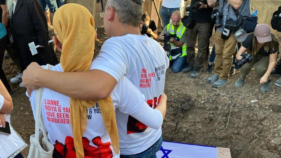 A man and woman stand by a graveside wearing t-shirts bearing the names of the Sharabi family