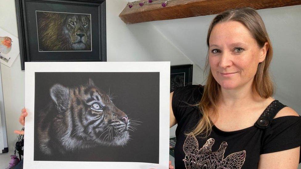 Smiling woman with long hair holds a mounted pencil drawing of a tiger