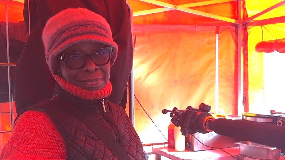 A woman stood behind her stall in a red jumper and hat and black jacket