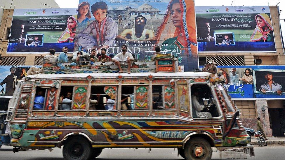 Pakistani men sit on a bus driving past a cinema displaying a billboard of the Indian and Pakistan joint venture movie 'Ramchand Pakistani' in Karachi.