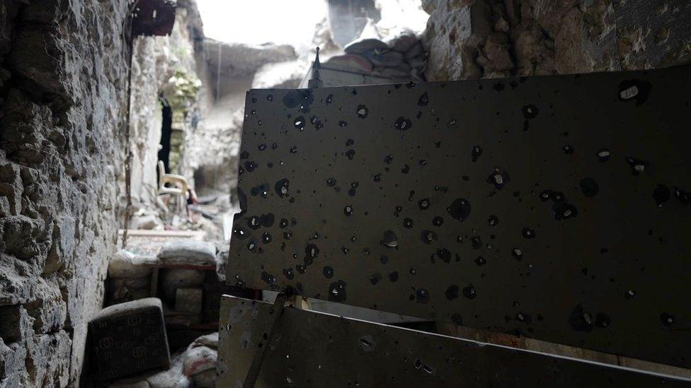 A sheet of metal with bullet holes, looking outside towards ruins