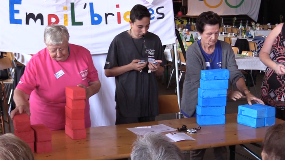 Two women stacking foam bricks in a tower as a judge stands between them with a timer and a whistle