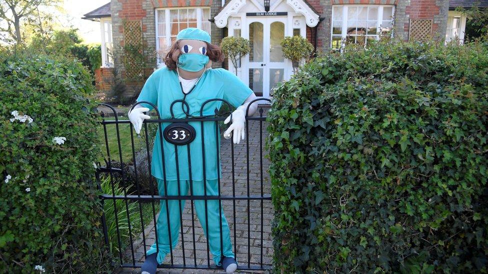 A figure representing a key worker is seen in a front garden as various scarecrows lighten the daily lockdown walk