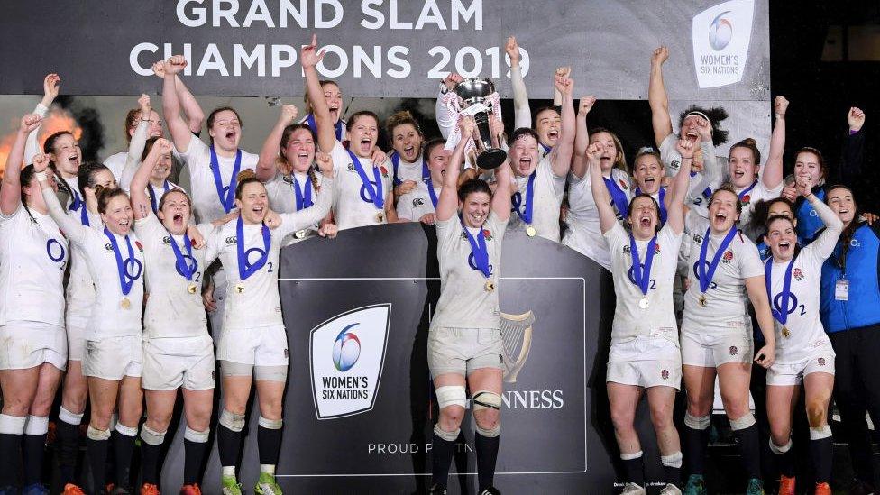 England women's rugby team wearing medals, celebrating with "grand slam champions" on wall behind