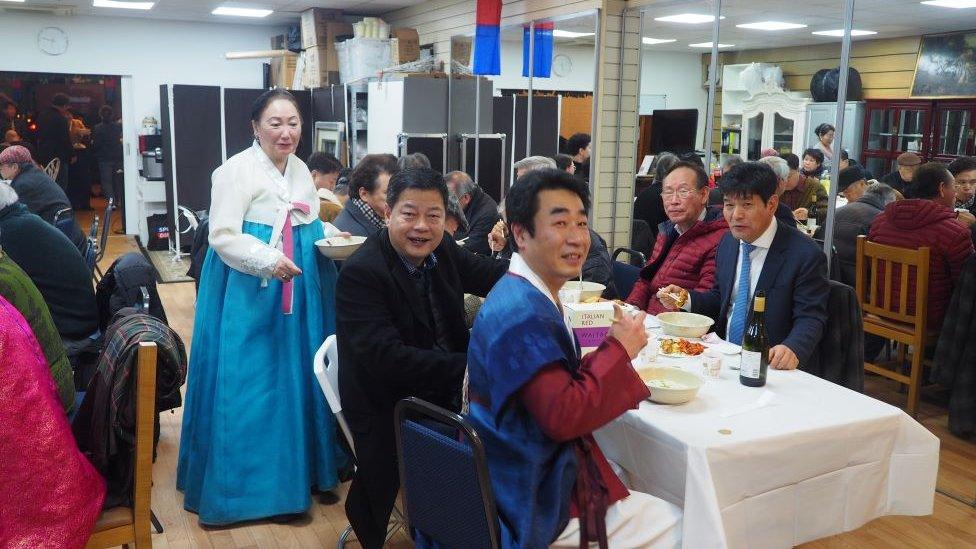 Woman in traditional Korean outfit stands at the top of a table. Men are seated at the table with food in front of them. They are all smiling at the camera.