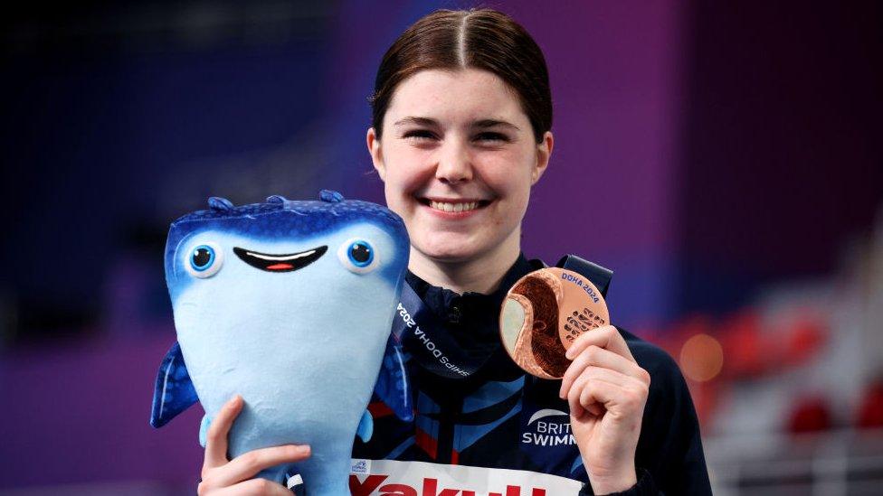 Bronze Medalist, Andrea Spendolini Sirieix of Team Great Britain pose with their medals during the Medal Ceremony after the Women's 10m Platform Final on day four of the Doha 2024 World Aquatics Championships. Andrea is a 19-year-old woman with dark brown hair tied back. She smiles at the camera as she holds up a bronze medal and a plush smiling stingray mascot. She wears a navy blue British Swimming tracksuit.