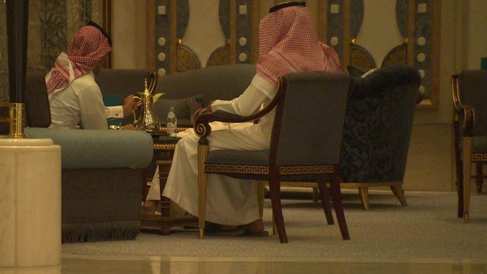 Saudi men in traditional dress sit in chairs in the Ritz Carlton in Riyadh, drinking coffee.