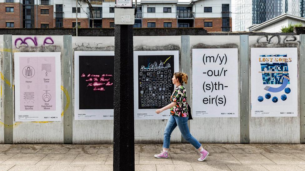 A woman walking past posters of artworks from the Poet Slash Artist exhibition