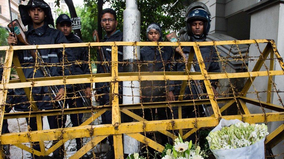 Bangladeshi policemen guard a check point where floral arrangements have been left by well-wishers on a road leading to an upscale cafe that was the site of a bloody siege in Dhaka, 3 July 2016