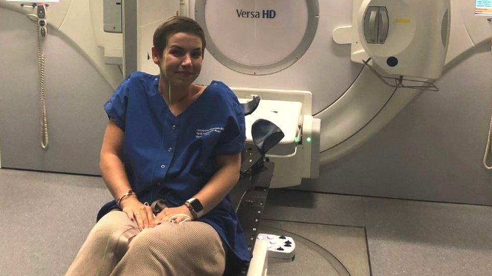 Danielle Moore in hospital near the radiotherapy equipment. She is wearing a blue hospital gown and is smiling at the camera.