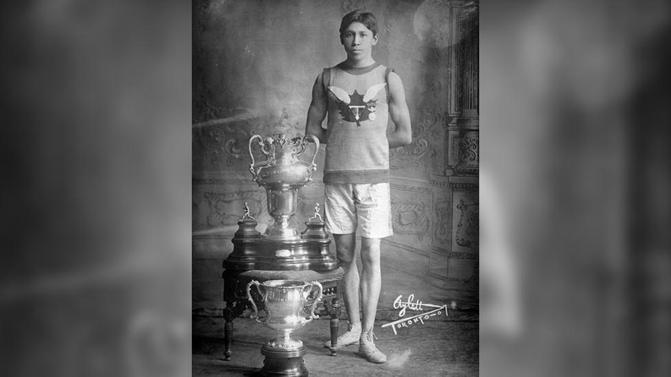 Tom Longboat posing with running trophies