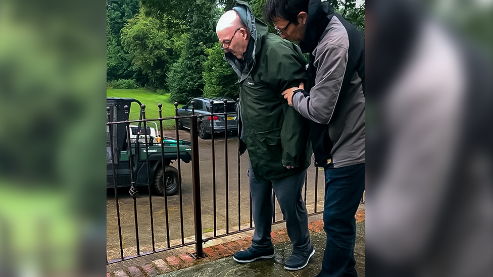 Roger walking outside holding a railing and being held on the other side by a physiotherapist